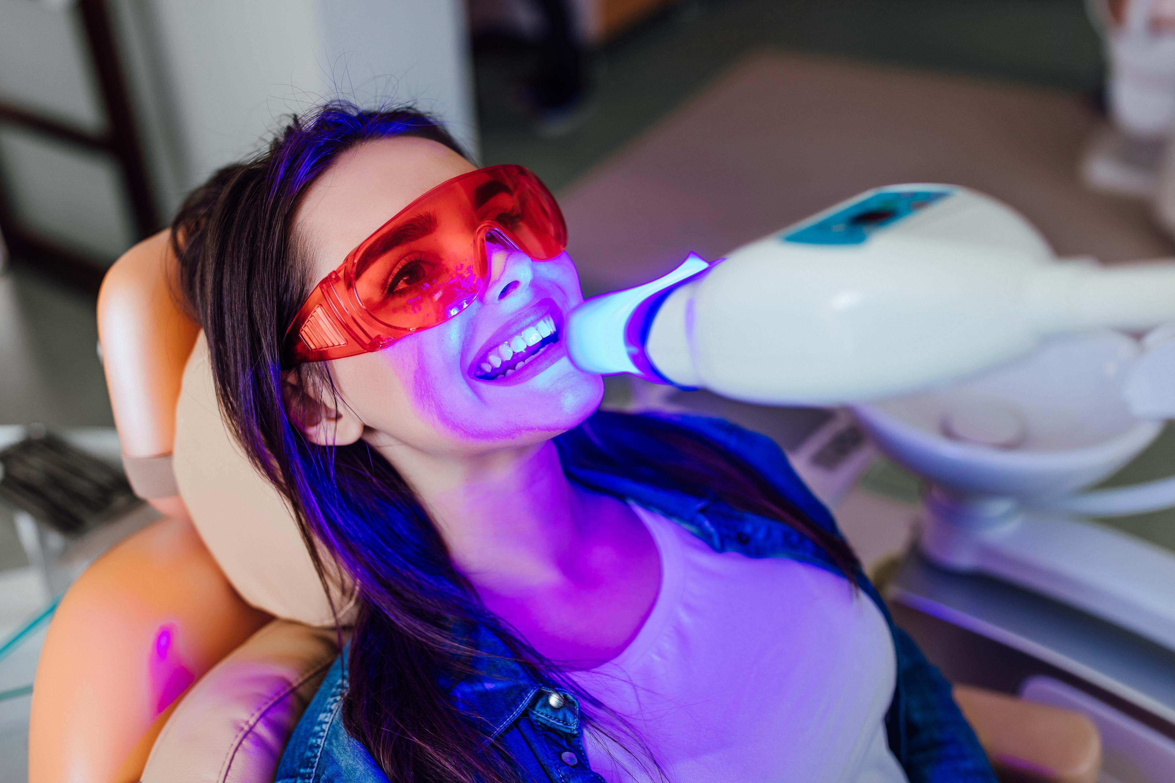 Woman wearing protective orange safety glasses reclines in a dental chair while receiving a professional teeth whitening treatment with a blue LED light device.